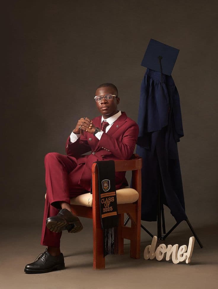 Graduate in a maroon suit seated beside a gown and mortarboard on a stand